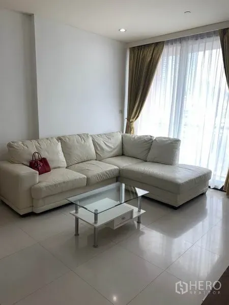 Condominium for rent in Khlong Toei, Bangkok - Sunlit corner with a white sectional sofa, glass coffee table and floor-to-ceiling curtains.