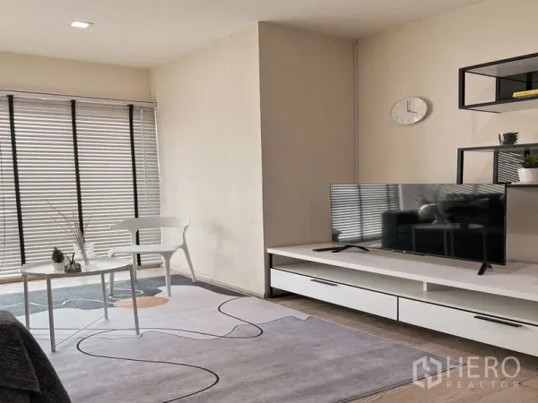Condominium for rent in Khlong Toei, Bangkok - Corner of living room featuring TV stand, patterned rug, large windows with blinds, chair and side table.