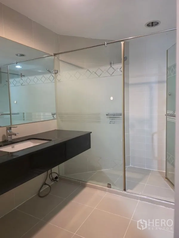 Condominium for rent in Huai Khwang, Bangkok - Vanity with large mirror beside a frosted glass shower in a tiled bathroom.