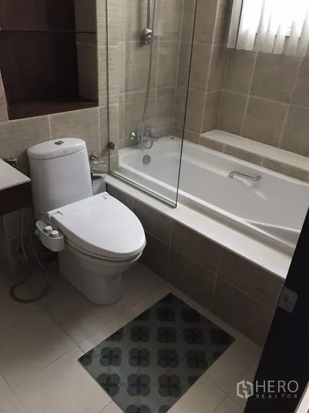 公寓 出租在Watthana, Bangkok - 浴室配浴缸玻璃隔断、手持花洒、坐便器与米色瓷砖。