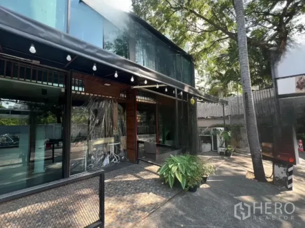 零售空间 出租在Watthana, Bangkok - 玻璃外立面侧视图，带遮荫露台、灯串、树木以及场内停车位。