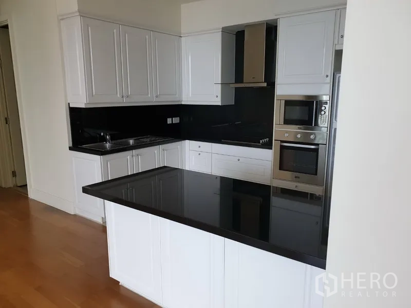 公寓 出租在Watthana, Bangkok - 现代L形厨房，白色橱柜与黑色石面台，内置烤箱、微波炉与炉灶。