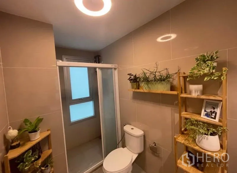 Condominium for sale in Bang Sue, Bangkok - Modern bathroom with glass shower enclosure, toilet and wooden plant shelving.