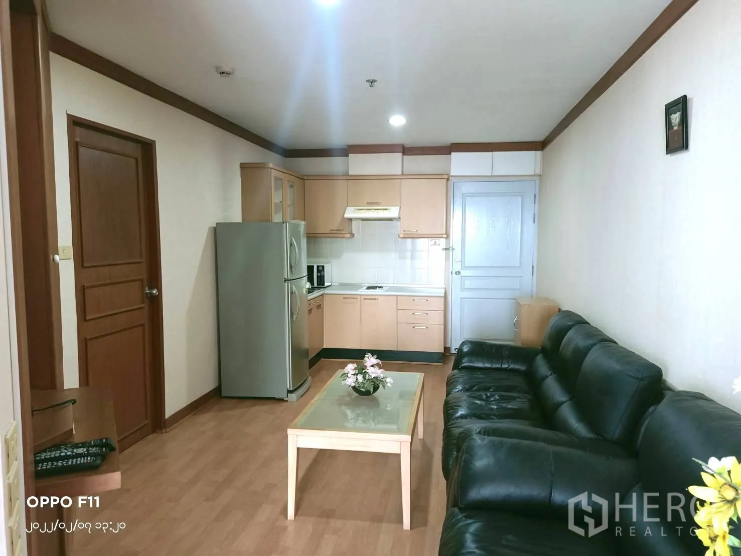 Condominium for rent in Khlong Toei, Bangkok - Open-plan living and kitchen with full-size refrigerator, black sofa and light wood cabinetry.