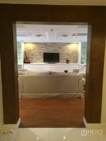 Condominium for rent in Khlong Toei, Bangkok - View into a spacious living room with beige sofas, a stone feature wall and TV console on warm wood flooring.