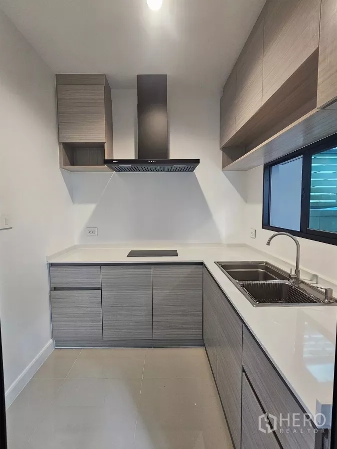 Townhouse for rent in Saphan Sung, Bangkok - Compact L-shaped kitchen with wood cabinets, cooktop, range hood and a window.