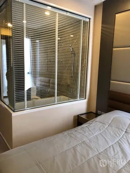 Condominium for rent in Watthana, Bangkok - Bedroom with padded headboard and a glass window looking into the bathroom with blinds.