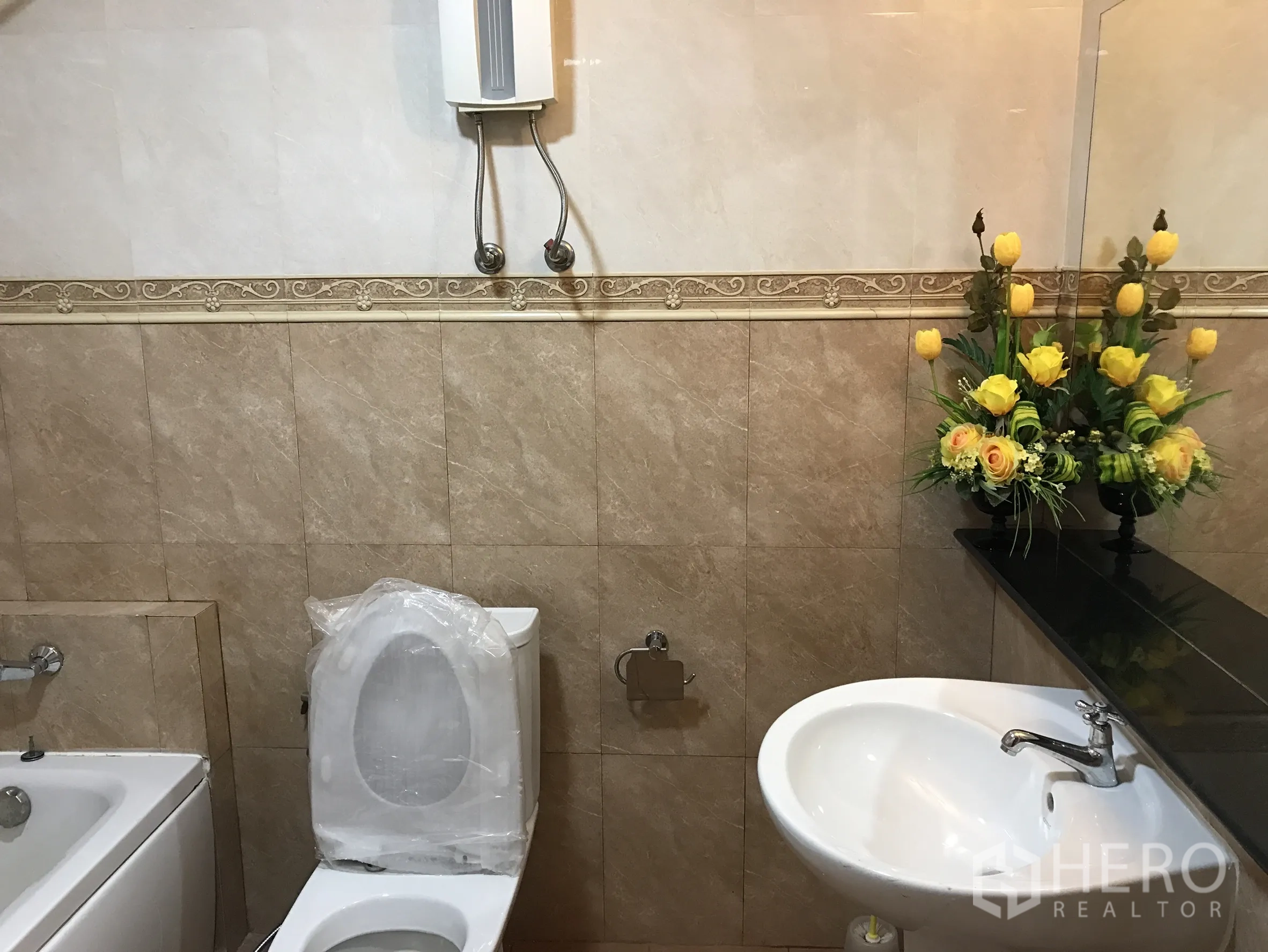 Condominium for rent in Khlong Toei, Bangkok - Tiled bathroom with bathtub, toilet, basin and yellow floral arrangement.
