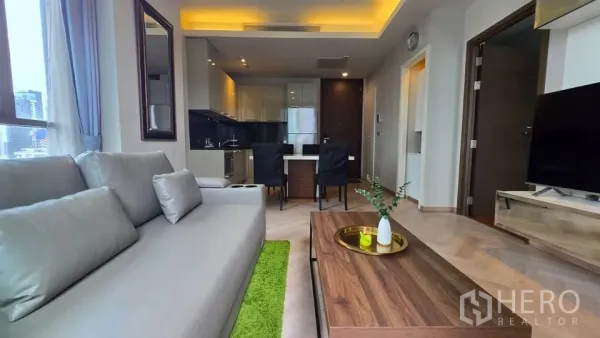 公寓 出租在Watthana, Bangkok - 采光充足的客厅，灰色沙发、木质茶几，开放式厨房与餐区，带灯槽天花。