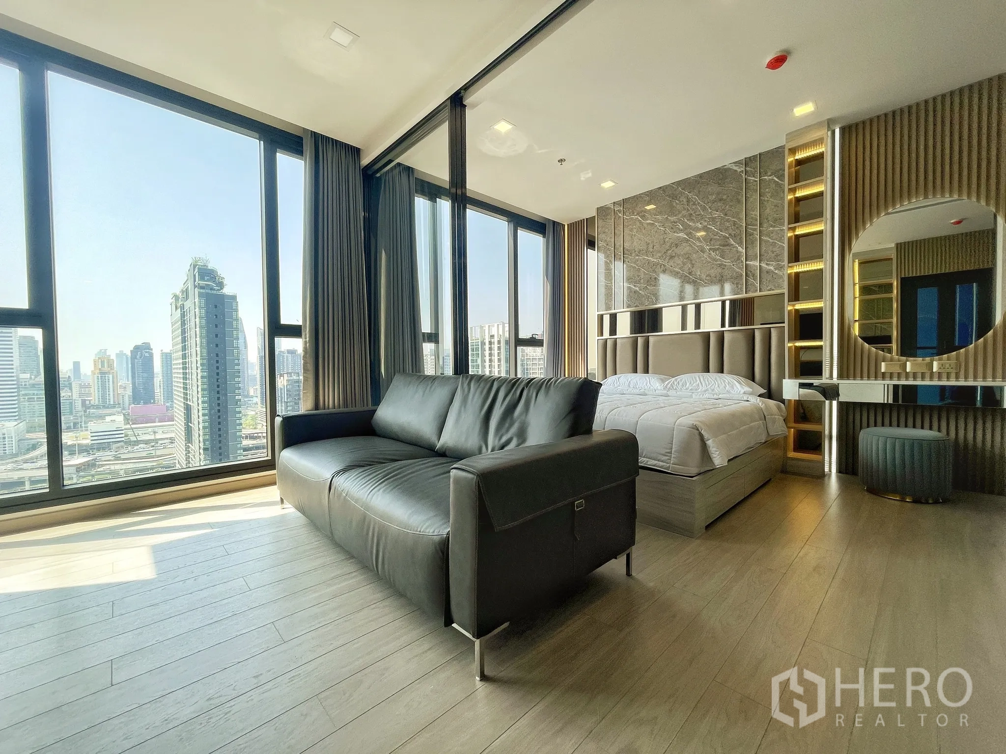 คอนโด for rent in Huai Khwang, Bangkok - พื้นที่นั่งเล่นและห้องนอนแบบเปิดโล่ง โซฟาหนังสีดำ เตียงนอน หน้าต่างกระจกสูงจรดเพดานมองเห็นวิวเมือง พร้อมโต๊ะเครื่องแป้งบิวท์อินและพื้นไม้