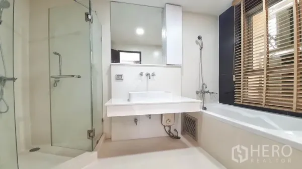 Condominium for rent in Khlong Toei, Bangkok - Primary bathroom with glass-enclosed shower, bathtub by the window with blinds, large mirror and vanity.