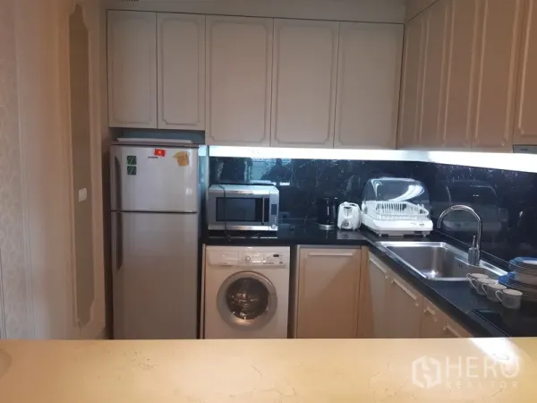 Condominium for rent in Khlong Toei, Bangkok - Compact L-shaped kitchen with white cabinets, black countertops, fridge, microwave, sink, and washing machine.