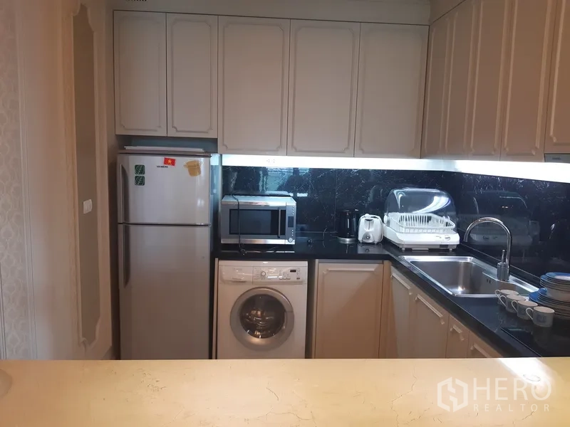 Condominium for rent in Khlong Toei, Bangkok - Compact L-shaped kitchen with white cabinets, black countertops, fridge, microwave, sink, and washing machine.