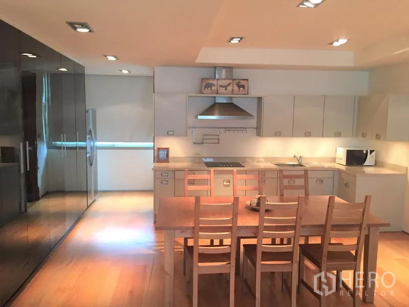 Condominium for rent in Khlong Toei, Bangkok - Contemporary kitchen with built-in cabinets, stainless range hood, full-size fridge and a wooden dining table.
