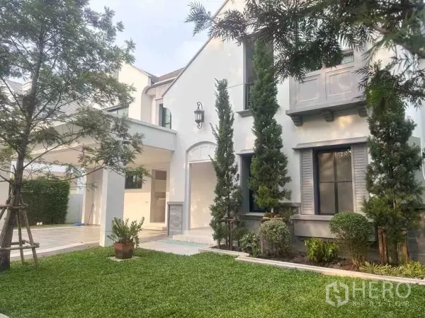 Detached House for rent or sale in Saphan Sung, Bangkok - Front lawn and covered carport beside modern white facade with tall evergreens