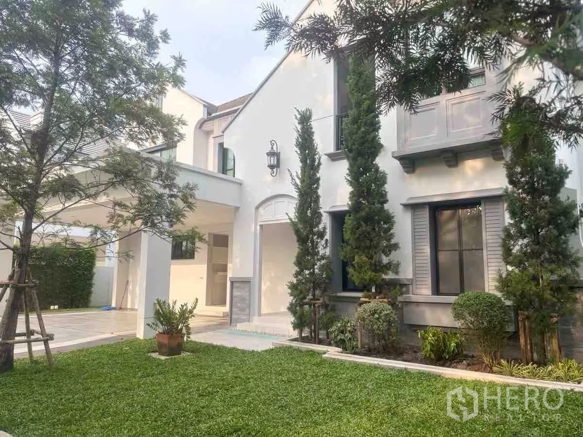 Detached House for rent or sale in Saphan Sung, Bangkok - Front lawn and covered carport beside modern white facade with tall evergreens