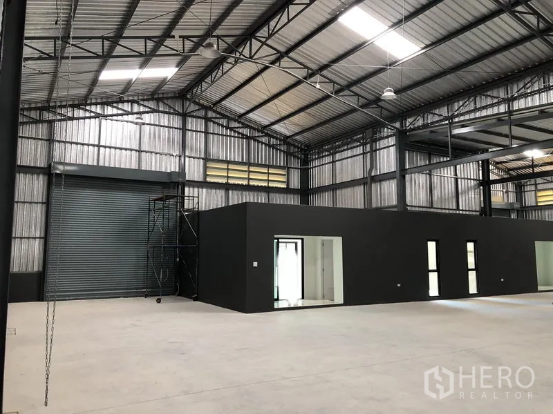 Factory for rent in Bang Yai, Nonthaburi - Interior view showing a roller shutter loading door beside dark grey office rooms under a high ceiling.