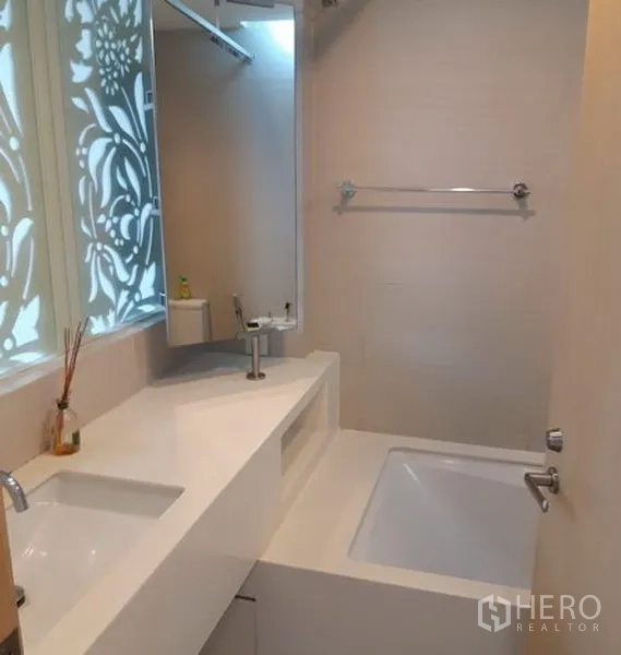 Condominium for rent in Khlong Toei, Bangkok - Modern bathroom featuring a long white vanity, under-mount sink, large mirror, built-in bathtub and decorative frosted window.