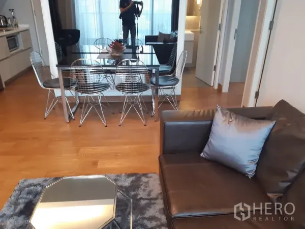 Condominium for rent in Khlong Toei, Bangkok - Open-plan living and dining area with glass table, mirror wall and warm wooden floors.