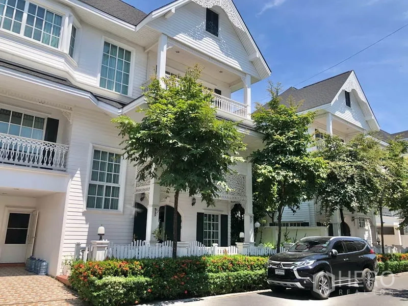 บ้าน ให้เช่าในMueang Samut Prakan, Samut Prakan - ด้านหน้าบ้านเดี่ยวสไตล์โคโลเนียลสีขาวหลายชั้น หลังคาจั่ว ระเบียง รั้วไม้พิกเกต ต้นไม้ และที่จอดรถ