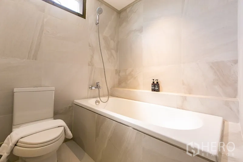 Apartment for rent in Khlong Toei, Bangkok - Bathroom with bathtub, handheld shower and light marble tiles.