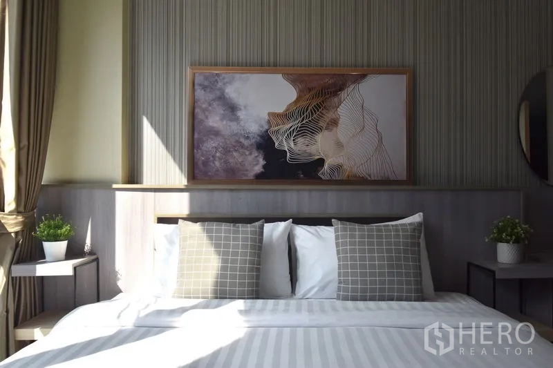 Condominium for rent in Khlong Toei, Bangkok - Sunlit bedroom with a large bed, patterned cushions, wall art, and bedside tables with plants.