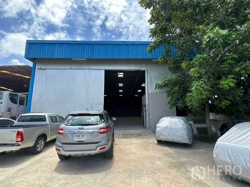 Warehouse for rent in Mueang Pathum Thani, Pathum Thani - Front exterior of metal-clad warehouse with blue roof trim, open loading door and parking area.