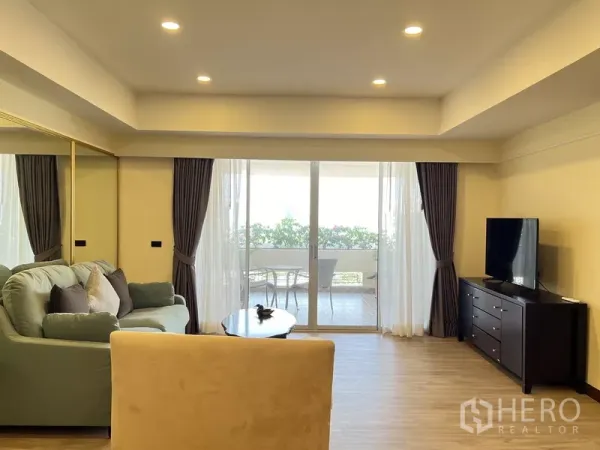 Condominium for rent in Khlong Toei, Bangkok - Bright living room with sofa, TV console and sliding glass doors opening to a leafy balcony with outdoor seating.