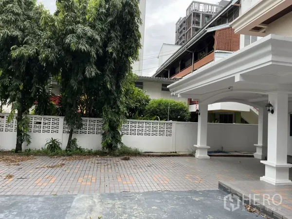 Detached House for rent in Watthana, Bangkok - Spacious front yard with tiled patio, white privacy wall and greenery beside the carport.