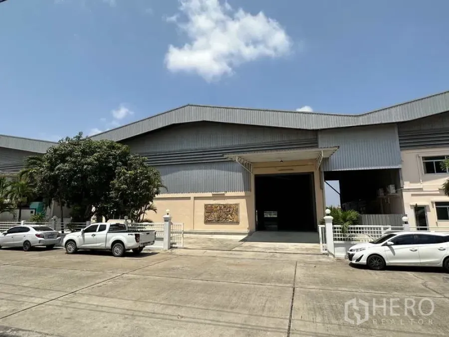โรงงาน ให้เช่า - มุมมองด้านหน้าของโรงงานผนังเมทัลชีท ประตูม้วนขนาดใหญ่ ทางเข้าแบบมีรั้วและที่จอดรถ ใต้ท้องฟ้าโปร่ง
