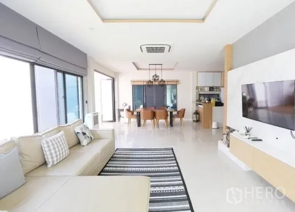 Detached House for rent or sale in Bang Kapi, Bangkok - Bright open-plan living and dining area with L-shaped sofa, wall-mounted TV and large windows.