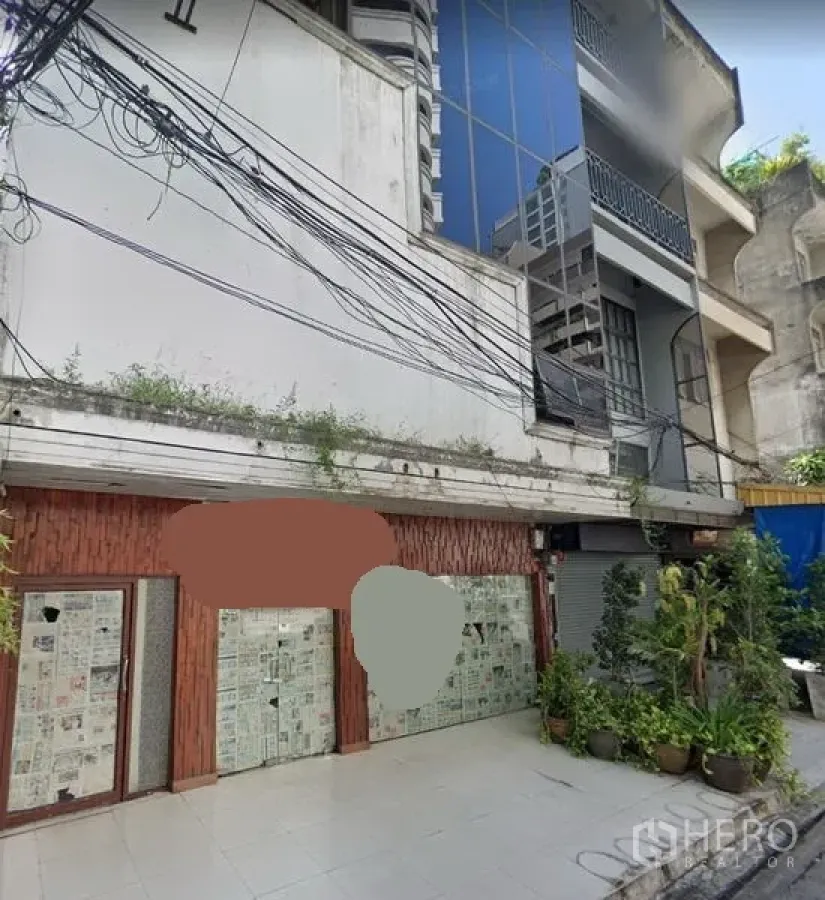 Shophouse for rent or sale in Khlong Toei, Bangkok - Street-facing shophouse exterior with tiled forecourt, glass storefronts covered with paper, potted plants, and overhead power lines in an urban Bangkok setting.