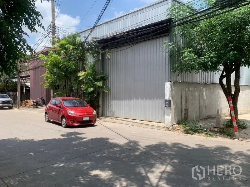 คลังสินค้า ให้เช่าในBang Kapi, Bangkok - ด้านหน้าโกดังผนังเมทัลชีท ประตูม้วนสูง ถนนเงียบ มีต้นไม้และรถจอด ในเขตบางกะปิ