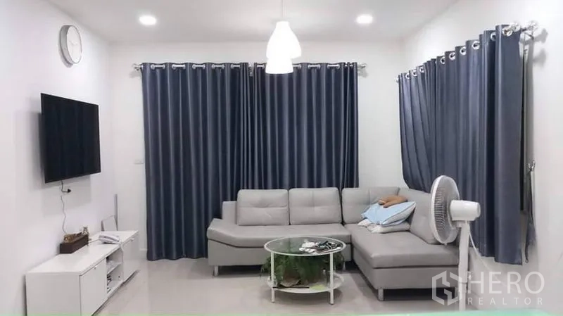 独立式住宅 出租或出售在Prawet, Bangkok - 客厅，L形沙发、壁挂电视与灰色窗帘