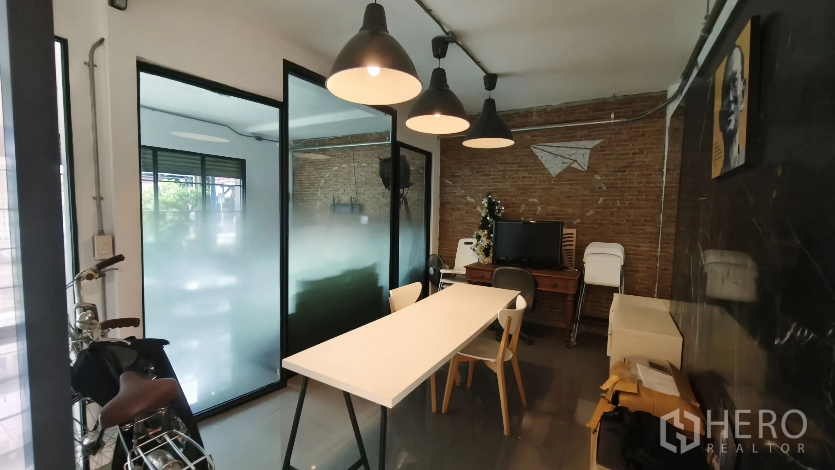 Shophouse for rent in Khlong Toei, Bangkok - Industrial-style workspace with long table, pendant lights, frosted glass partitions, and an exposed brick wall.