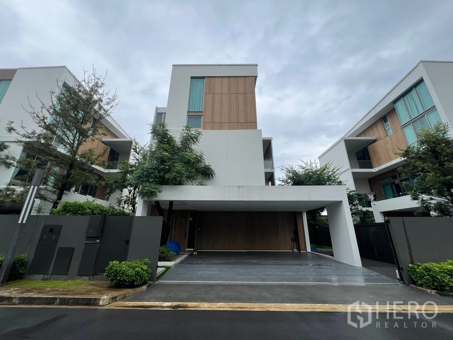 Detached House for rent in Saphan Sung, Bangkok - Front elevation of a modern three-storey detached house with carport and wood accents on a cloudy day.