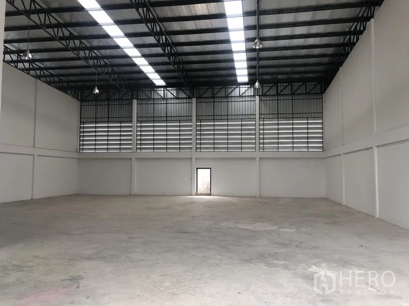 Factory for rent in Sai Noi, Nonthaburi - Large warehouse hall with high steel truss roof, skylight panels and a rear access door.