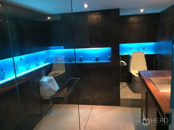 Condominium for rent in Khlong Toei, Bangkok - Modern bathroom with dark tiles, glass partition and blue backlit display niches.