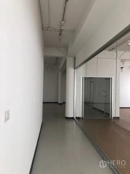 Office Space for rent in Bang Rak, Bangkok - Bright corridor with white walls, high ceiling and a glass partition leading to a wood-floor workspace.