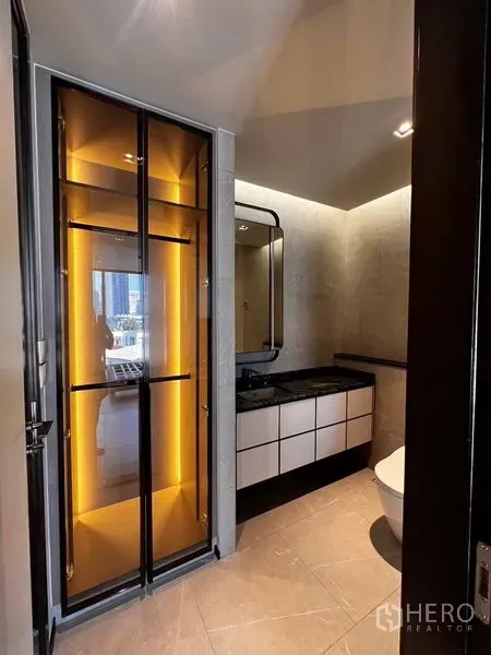 Condominium for sale in Watthana, Bangkok - Modern bathroom with backlit glass cabinet, black stone vanity, wall mirror and toilet under warm lighting.