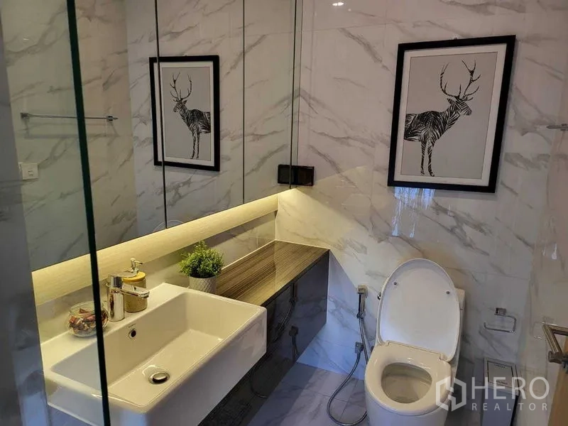 Condominium for rent in Watthana, Bangkok - Modern bathroom with marble‑effect tiles, large vanity with backlit mirror and toilet.