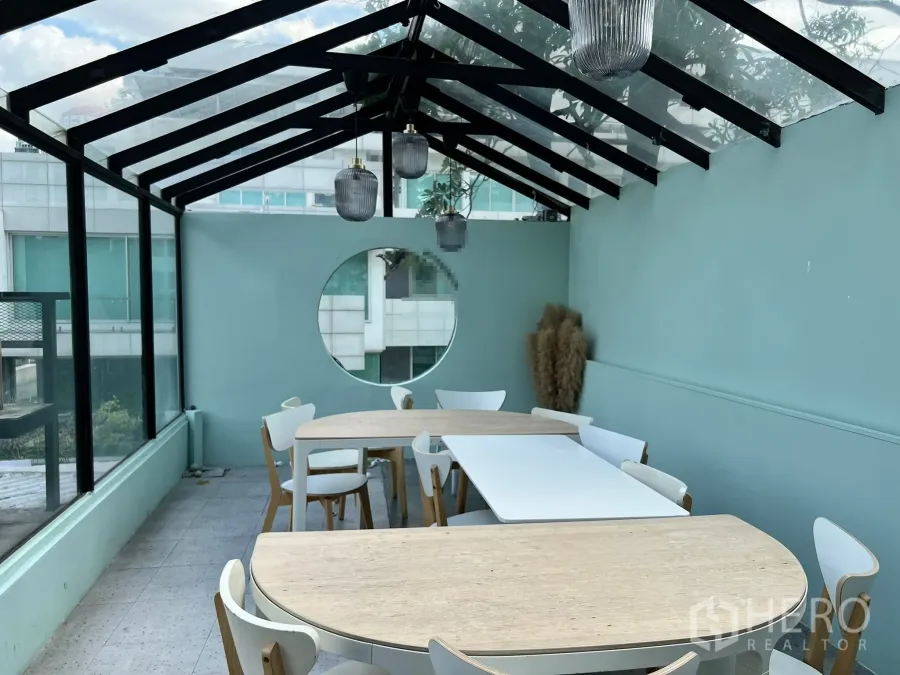 零售空间 出租在Watthana, Bangkok - 玻璃屋顶的温室用餐区，木质桌椅、薄荷绿墙、圆形窗与吊灯。