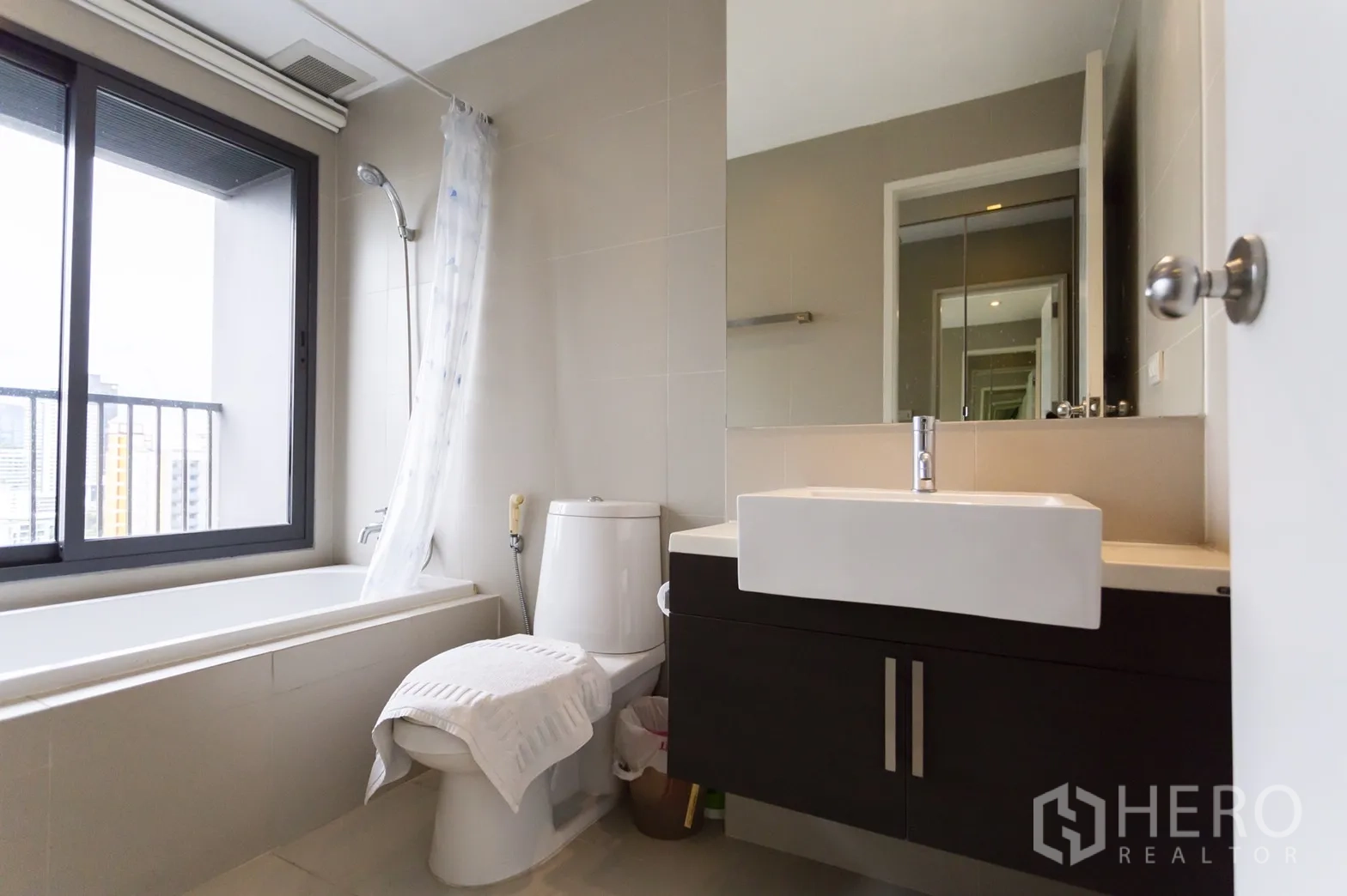 Condominium for rent in Khlong Toei, Bangkok - Contemporary bathroom featuring a bathtub by a large window, toilet and square-basin vanity.