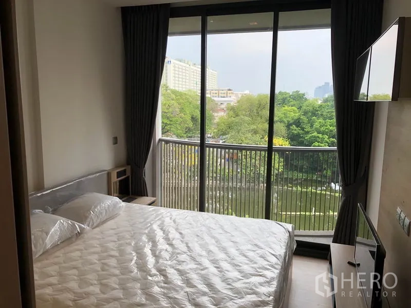 Condominium for rent in Watthana, Bangkok - Bedroom with double bed opening to a balcony with treetop and canal views, wall-mounted TV and blackout curtains.