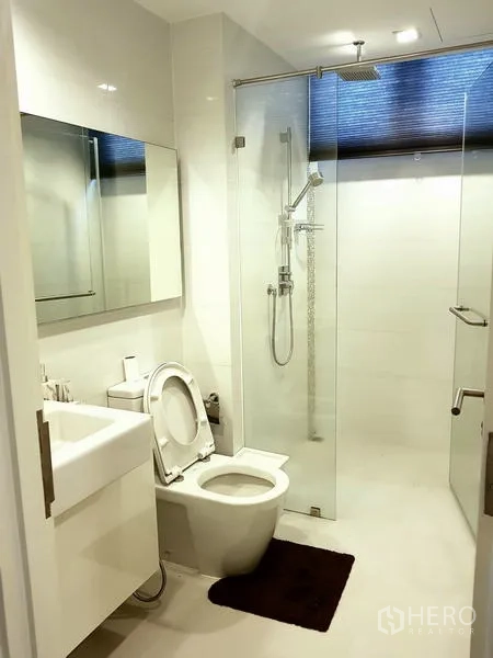 Condominium for rent in Sathon, Bangkok - Second bathroom with glass‑enclosed rain shower, vanity and toilet in white tones.