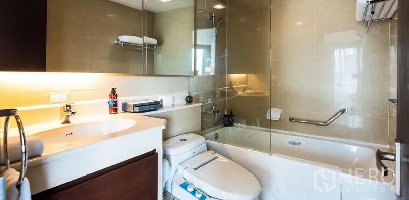 Condominium for rent in Khlong Toei, Bangkok - Contemporary bathroom featuring a glass-screened bathtub, large mirror and warm lighting.