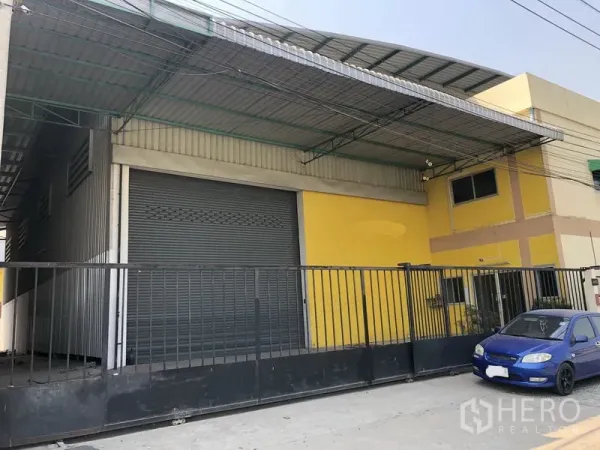 Factory for rent in Pathum Thani - Front exterior of a yellow factory with a large roller shutter door, covered loading canopy, gated entrance and a parked blue car.