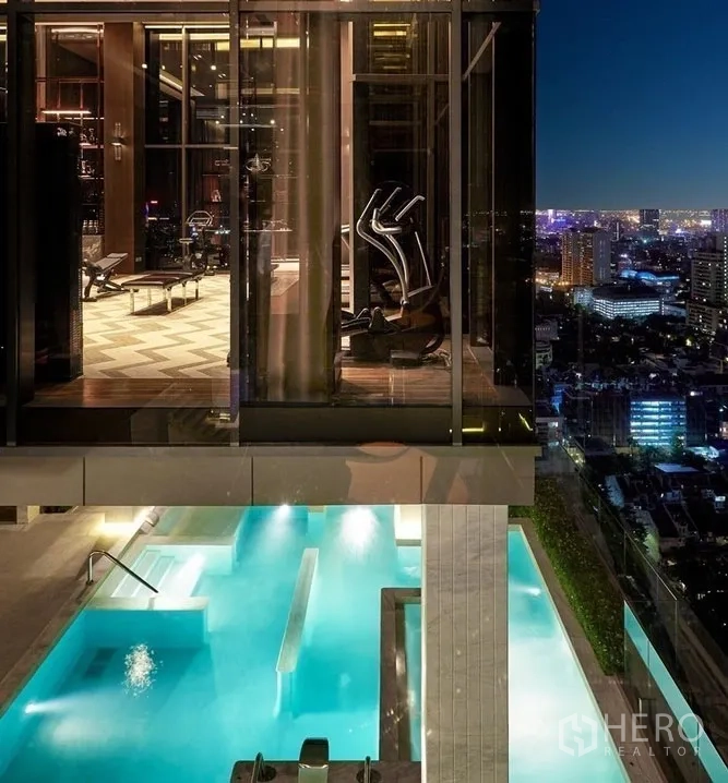 Condominium for sale in Watthana, Bangkok - Glass-walled fitness center overlooking the rooftop pool and Bangkok skyline at night.