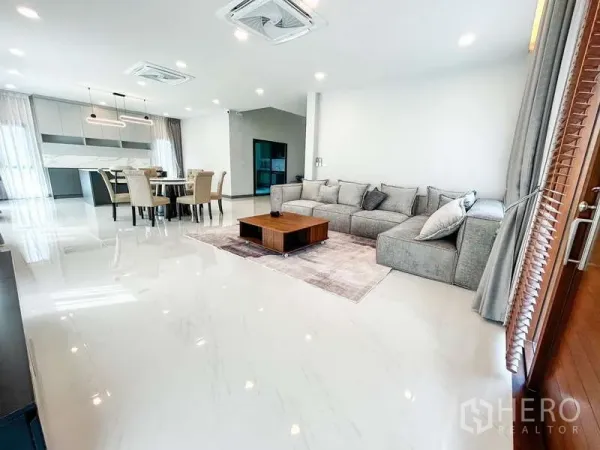 Detached House for rent in Bang Phli, Samut Prakan - Open-plan living and dining area with large grey sectional sofa, coffee table and shiny white floors.