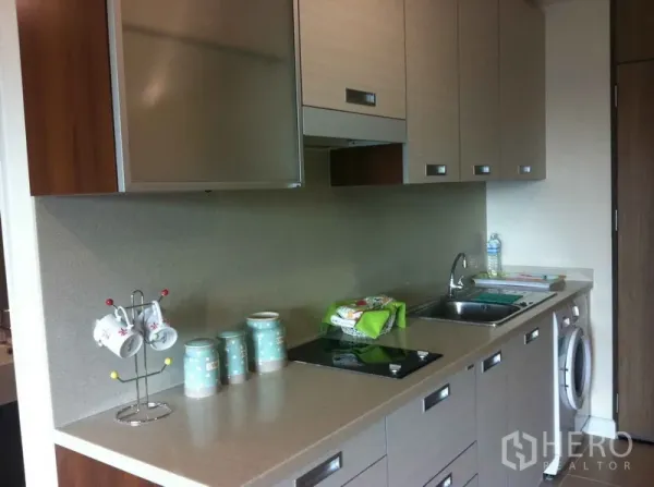 Condominium for rent in Khlong Toei, Bangkok - Modern galley kitchenette with upper cabinets, electric hob, sink and in-unit washer.
